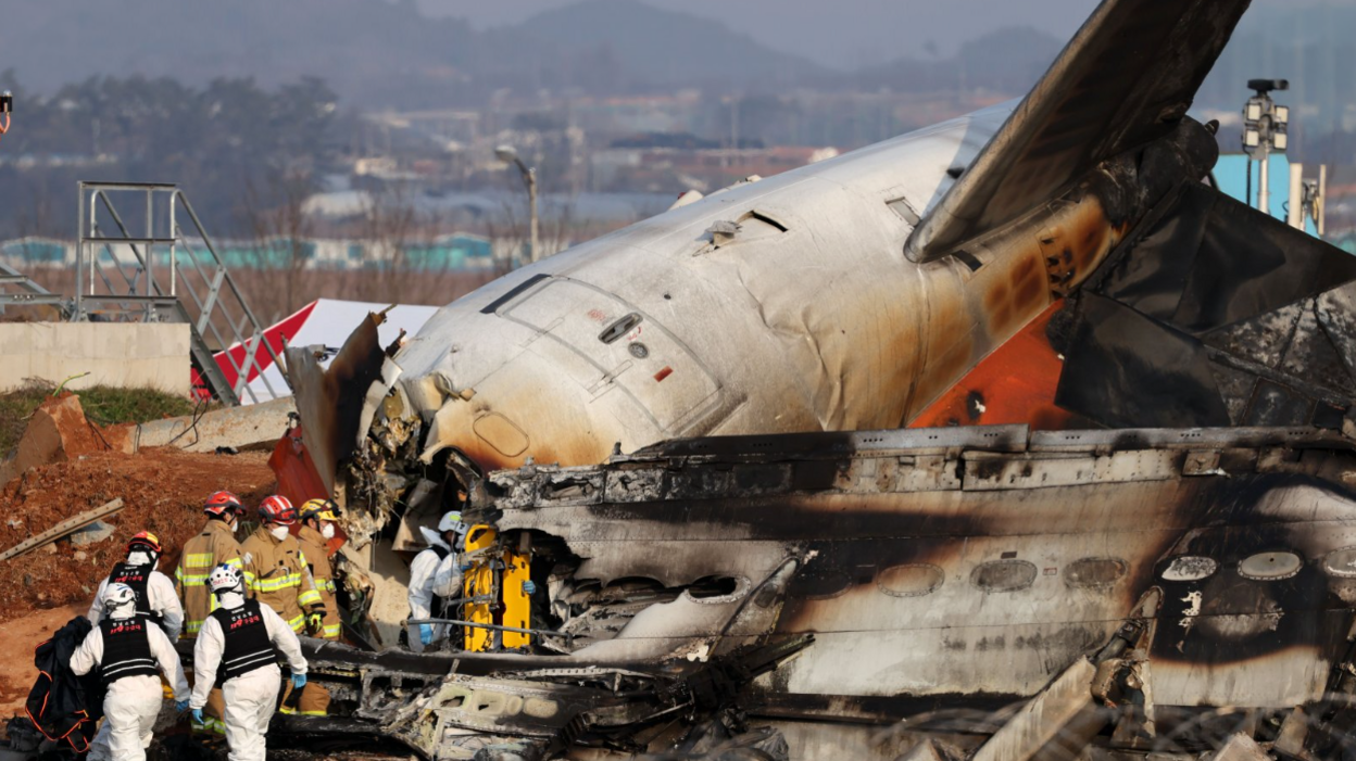 Firefighters and rescue workers in suits and helmets approach the charred wreckage of the plane. 