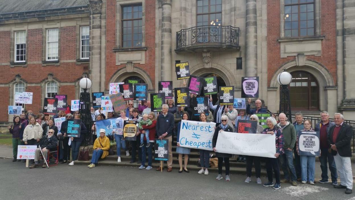 A group of people standing outside a large building, holding colourful banners. Some banners say 'bring catchment back' and 'save our schools'.
