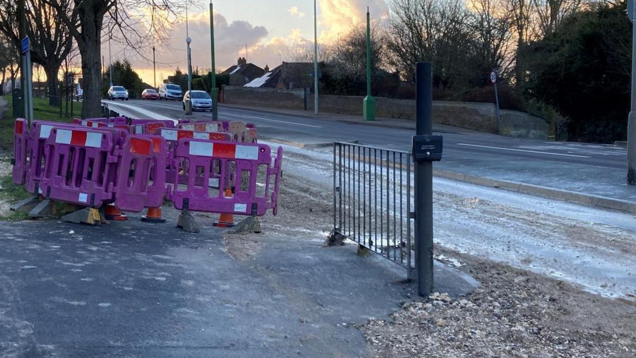 Purple temporary fencing around a hole by the side of Dereham Road. Gravel is strewn across the road and path around the hole. Cars are travelling on the opposite carriageway.