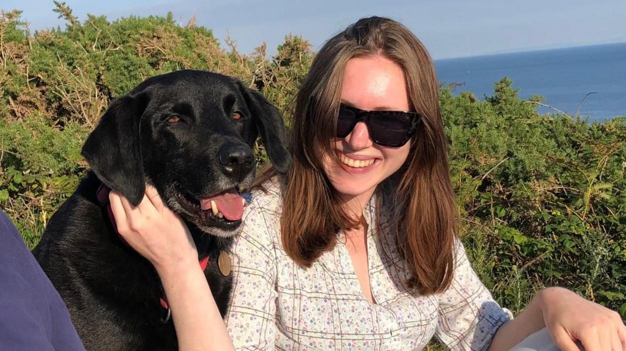 Rhiannon has long dark hair and is wearing a patterned white top and sunglasses. She is sat next to the family's black labrador, Jet, near Langland in the summer of 2022.