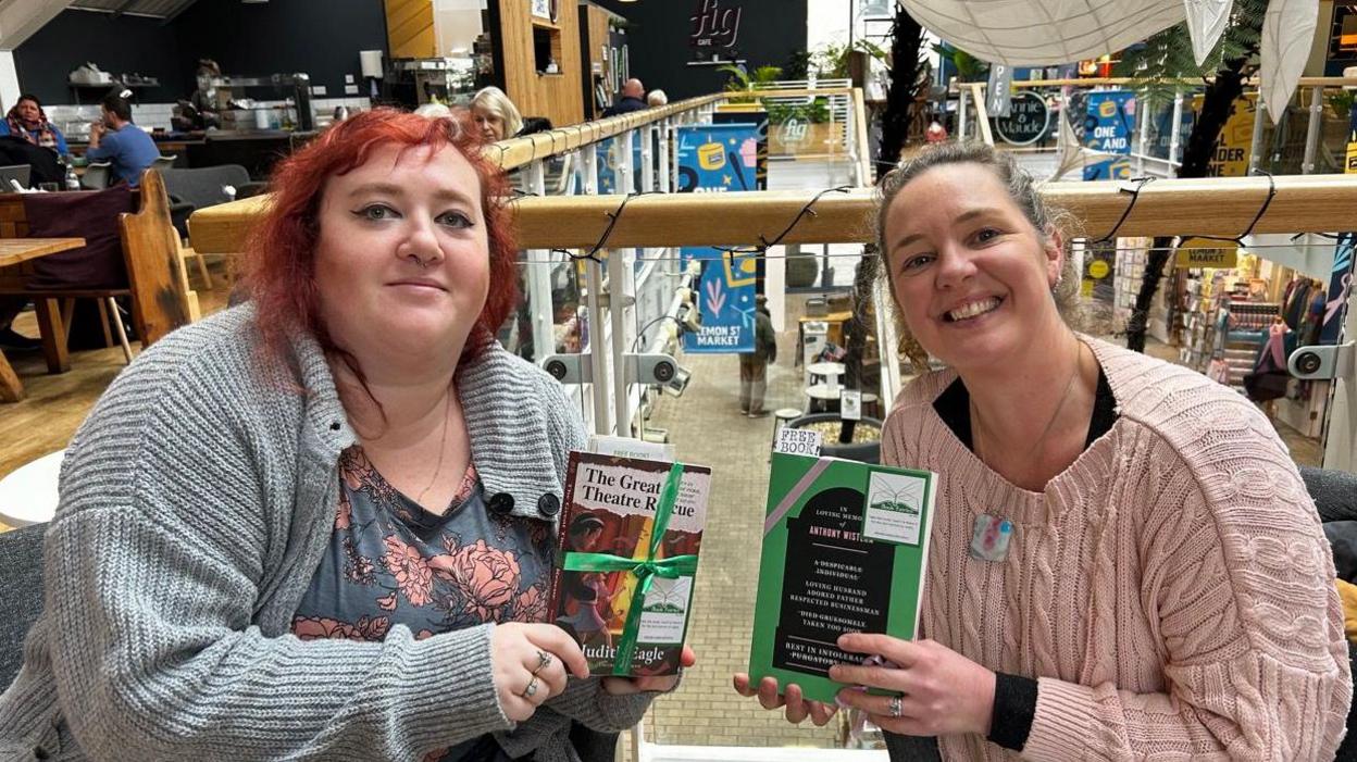 Carys Wiggans, on the left, with red hair, wearing a grey cardigan and a blue top with roses on it. She has a ring on her middle finger and her ring finger. She is Holding a book, titled The Great Theatre Rescue, which is wrapped in green ribbon and has a book fairy sticker on it. Kate Pearson, on the right, wearing a pink jumper, her blonde hair is pulled back. She is wearing a necklace, the pendent is square and is multi-coloured with white, pink and blue. She is wearing a ring on her ring finger. She is holding a green book, with pink ribbon on it and a book fairy sticker, with a bookmark which says 'Free Book' sticking out of the top. 