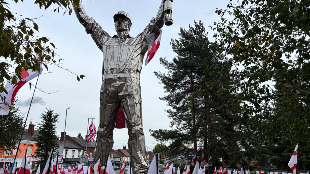 A silver statue of a man wearing a hard hat and holding a lamp is standing on a roundabout with the red and white cross of St George's flags at its feet. There are trees and shops in the background.
