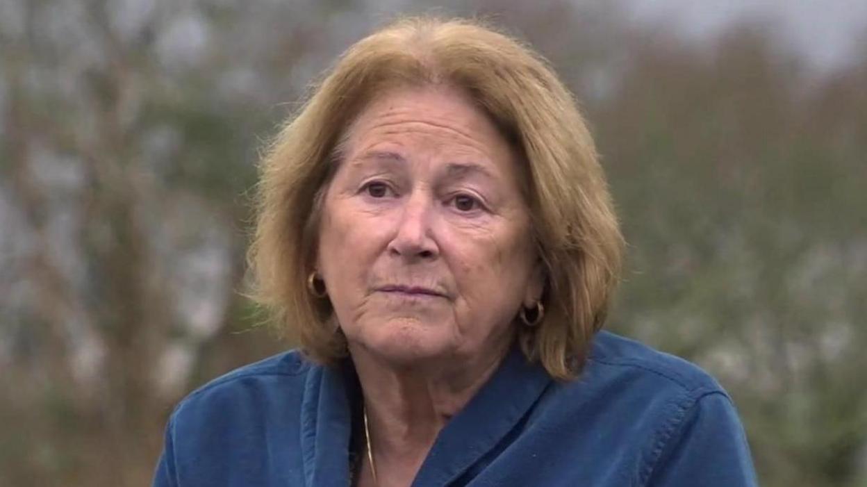 Linda Taylor, an older woman with shoulder-length light brown hair. She is wearing a blue jacket. There are some trees in the background.