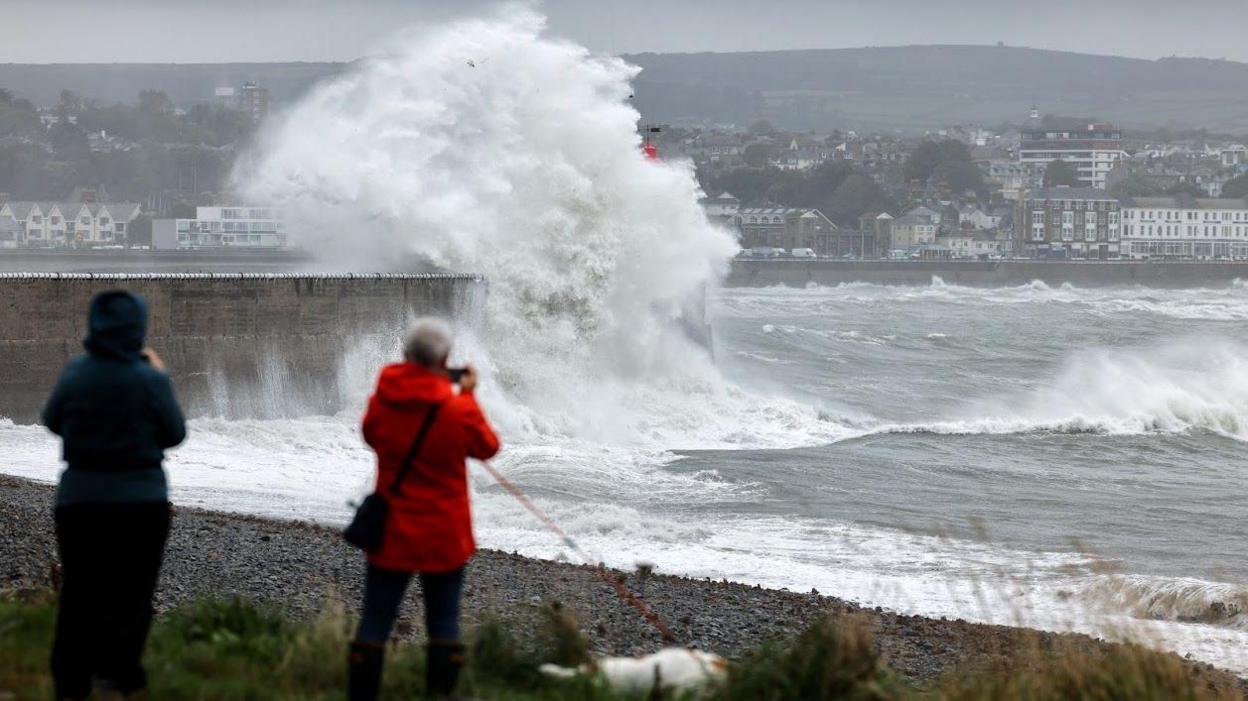 A foamy wave splashes over the lighthouse at Newlyn harbour, houses are seen in the background while the stones of a beach pepper the foreground. A man and a woman in a red coat are taking photos of the scene