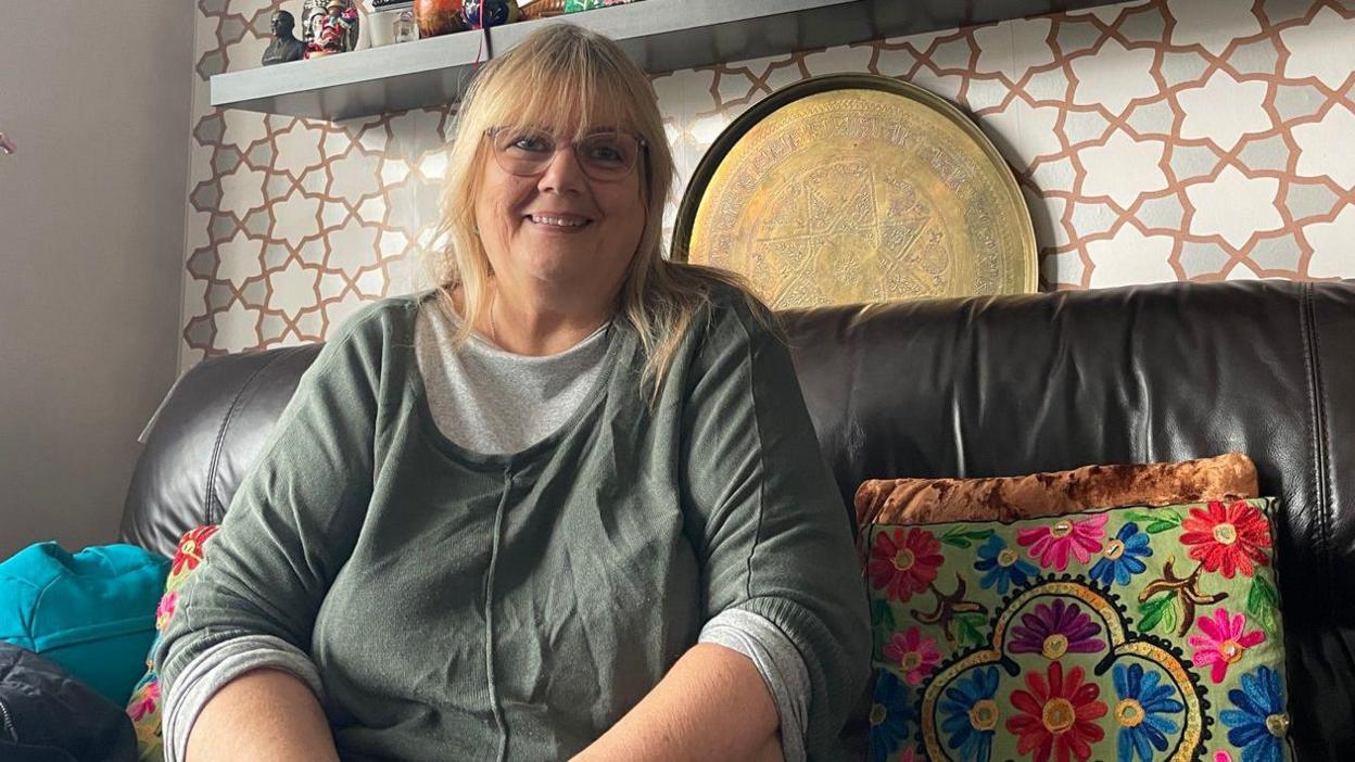 A blond lady with glasses and a grey long-sleeved shirt. She is sitting on a brown sofa and smiling. 