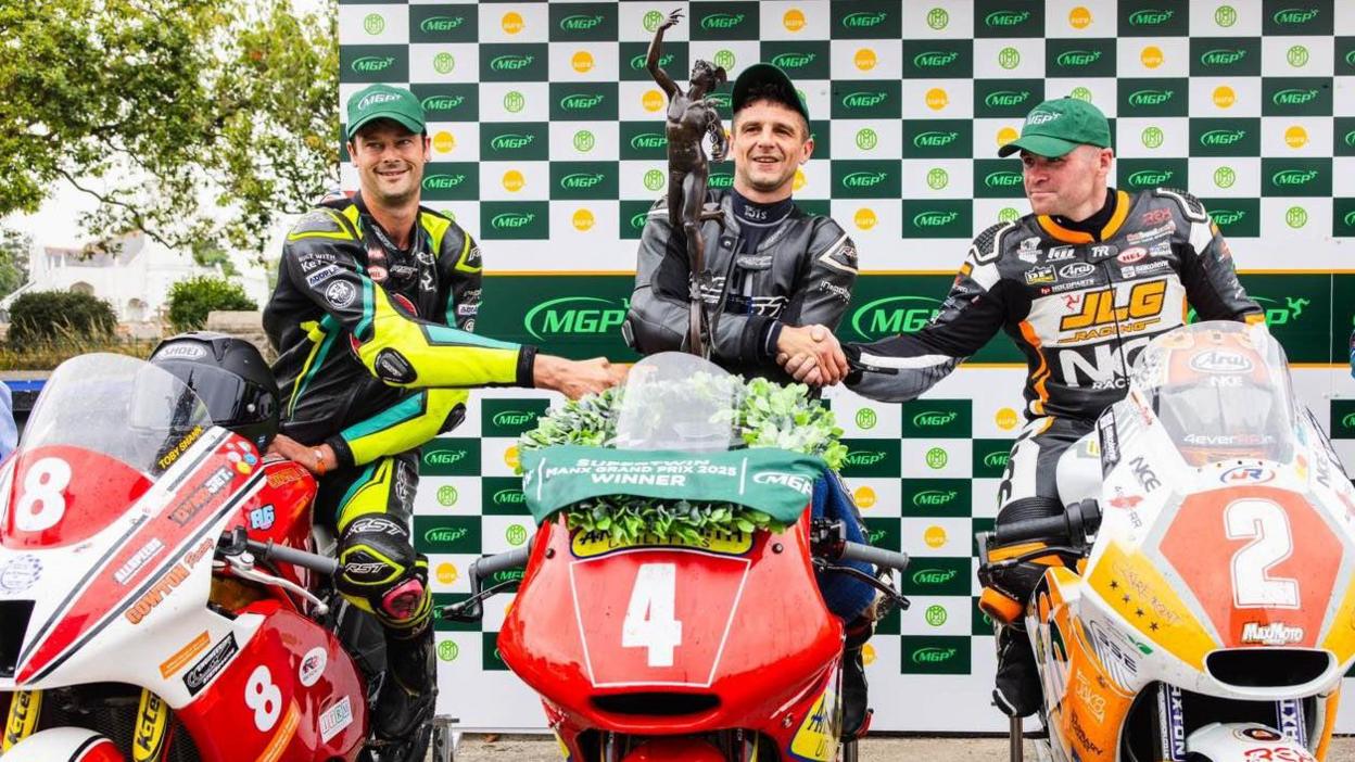 The top three riders on their bikes shaking hands. Lewis Arrowsmith is holding the Supertwin trophy and has a laurel garland on his bike.