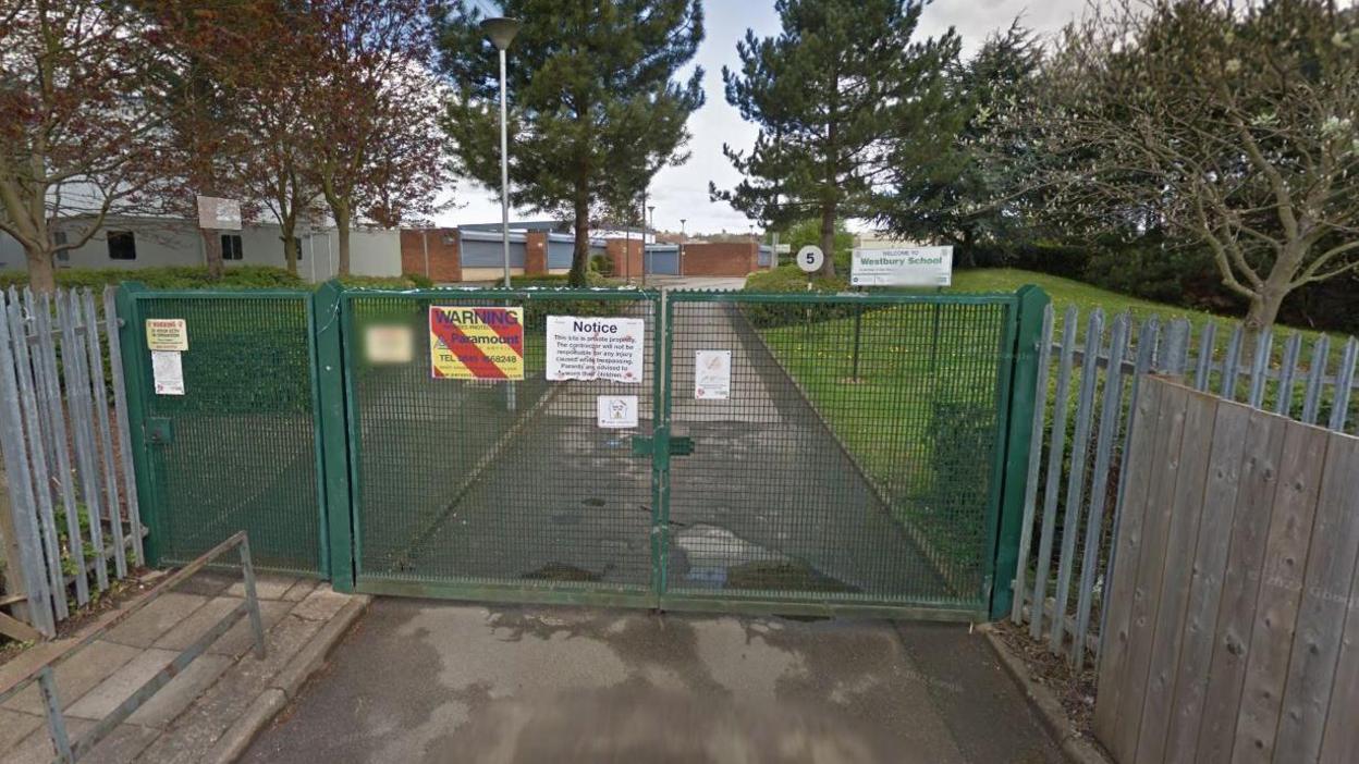 A Google image of the front of Westbury Academy, in Bilborough, Nottinghamshire. The photograph shows a green gate and behind it is a sign saying "Westbury School" and the school building 