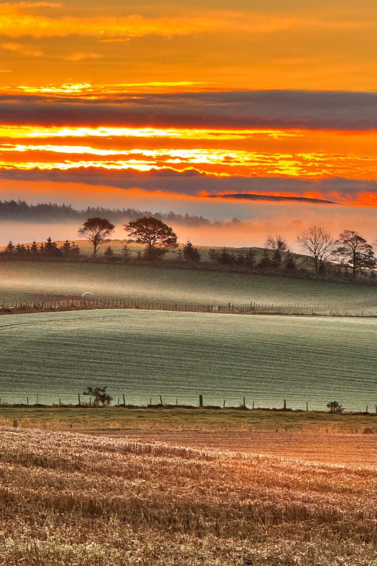 The sunset is orange and red in colour. There is farmland and trees and a mist drifts across the landscape.