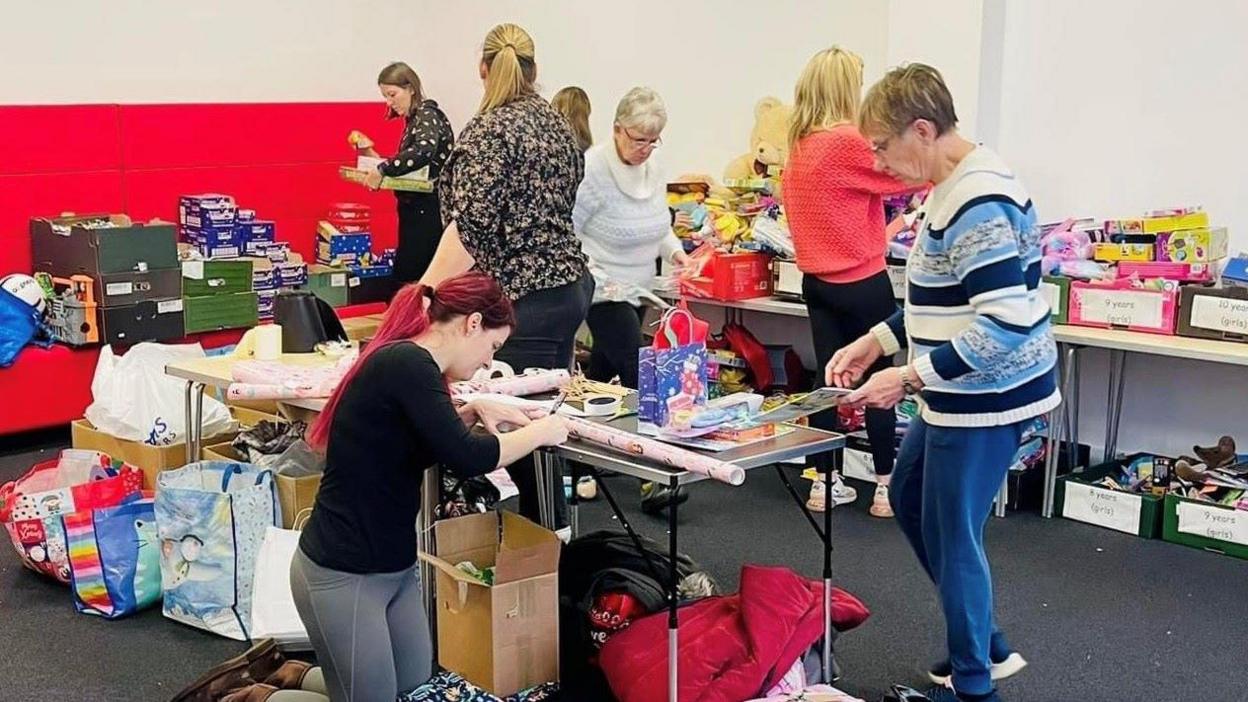 Seven people are scattered around a room - mostly stood at gift tables. In the foreground is a woman kneeling down using wrapping paper, while to her right is another woman looking at the table. Behind them is a group looking through gifts with their backs to the camera. There are lots of boxes of toys and chocolates.