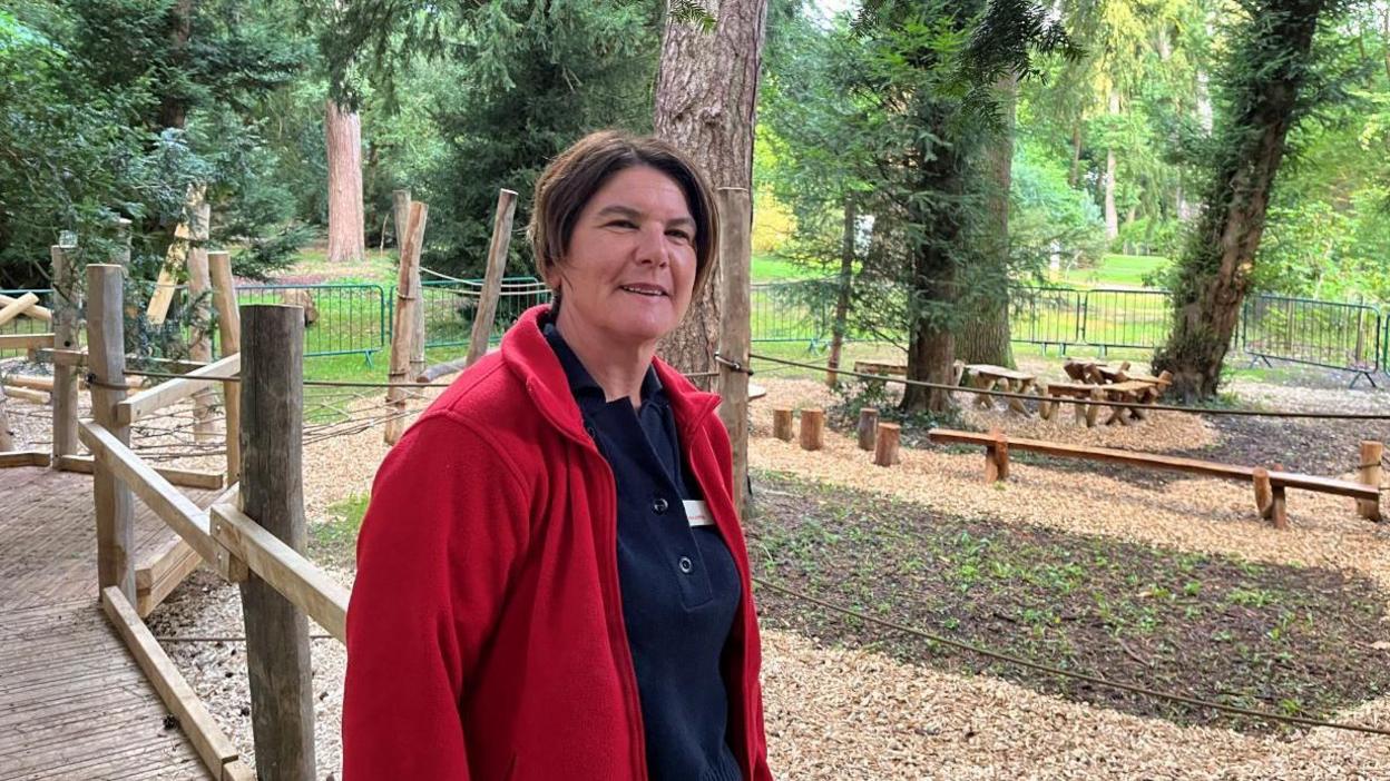  Emma Griffiths wearing a black polo shirt and a red fleece zip up jacket. She has short brown hair and a name tag on. In the background there are trees, bark chipping paths and wooden climbing frames 