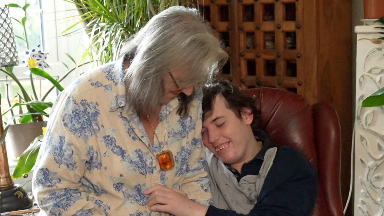 Chantal Chaervey rests on the arm of red leather chair which her son Harry sits in. She has shoulder length grey hair and wears a white blouse with blue flowers on it. Harry smiles as he rests his head on his mother's shoulder. He has dark hair and wears a grey and blue long sleeved top. 