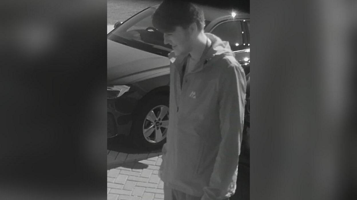 Ellis Cox is seen smiling as he stands in the driveway of his aunt's house in this black and white image. He is wearing a zipped raincoat and trousers. A car can be seen just to his right.