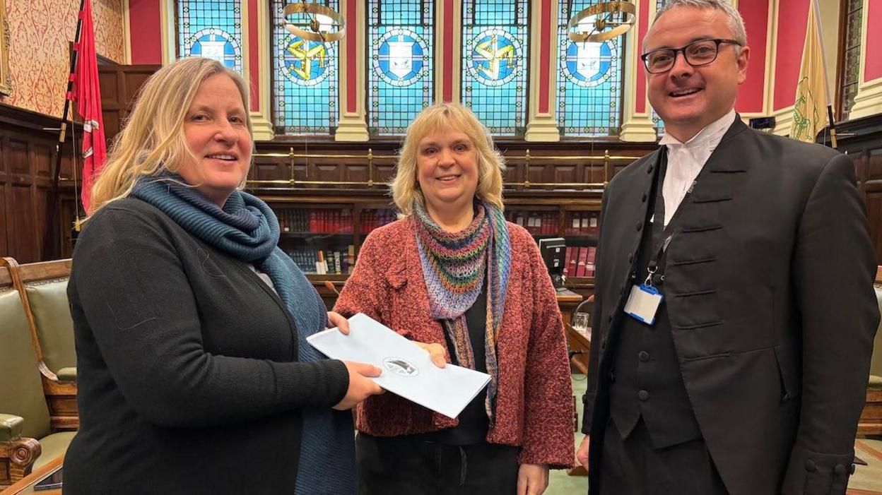 Christa-Marion Vhiohl, who is wearing a pink cardigan and colourful scarf and has blonde hair, standing in between Michelle Haywood, who also has blonde hair, and Juan Watterson, who has grey hair and is wearing a suit, to present a petition in the House of Keys chamber.