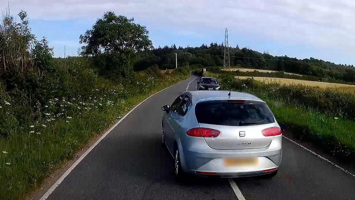 A dashcam image showing a silver car dangerously overtaking another vehicle on a country road as another car approaches in the opposite direction.