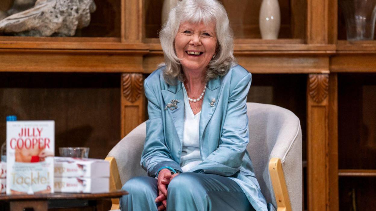 Jilly Cooper, a woman with grey shoulder length hair wearing a satin blue suit sitting in a chair with a pile of her books next to her. The books say 'Tackle' on them.