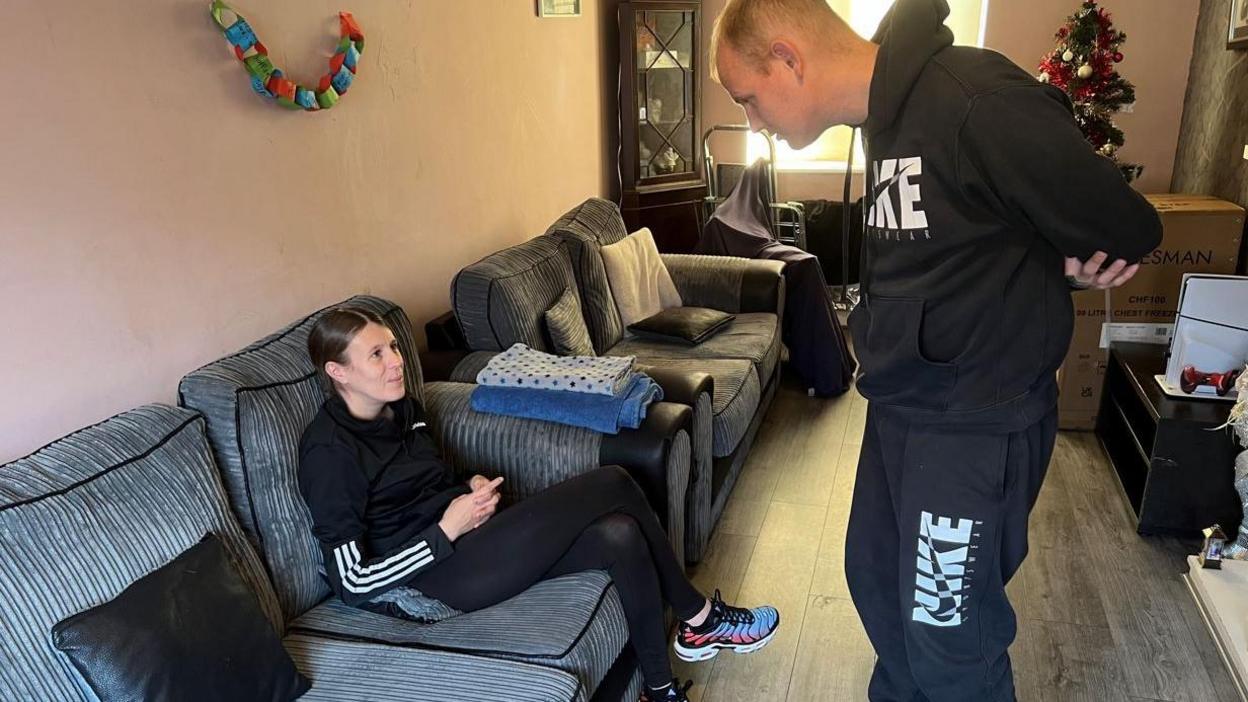 A woman wearing black sportswear and trainers sits on a grey sofa while Mr Johnson stands in the living room nearby