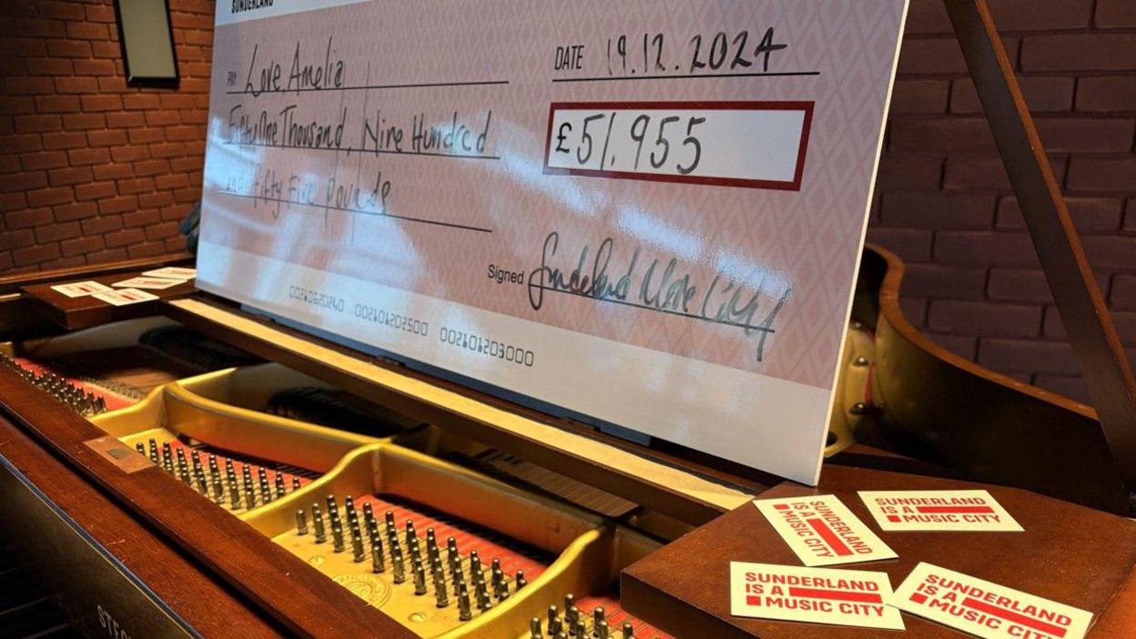 A large novelty check valued at £51,955 made out to Love, Amelia from Sunderland Music City. It is dated 19 December 2024. It is standing on a grand piano, which also holds flyers for Sunderland Music City.