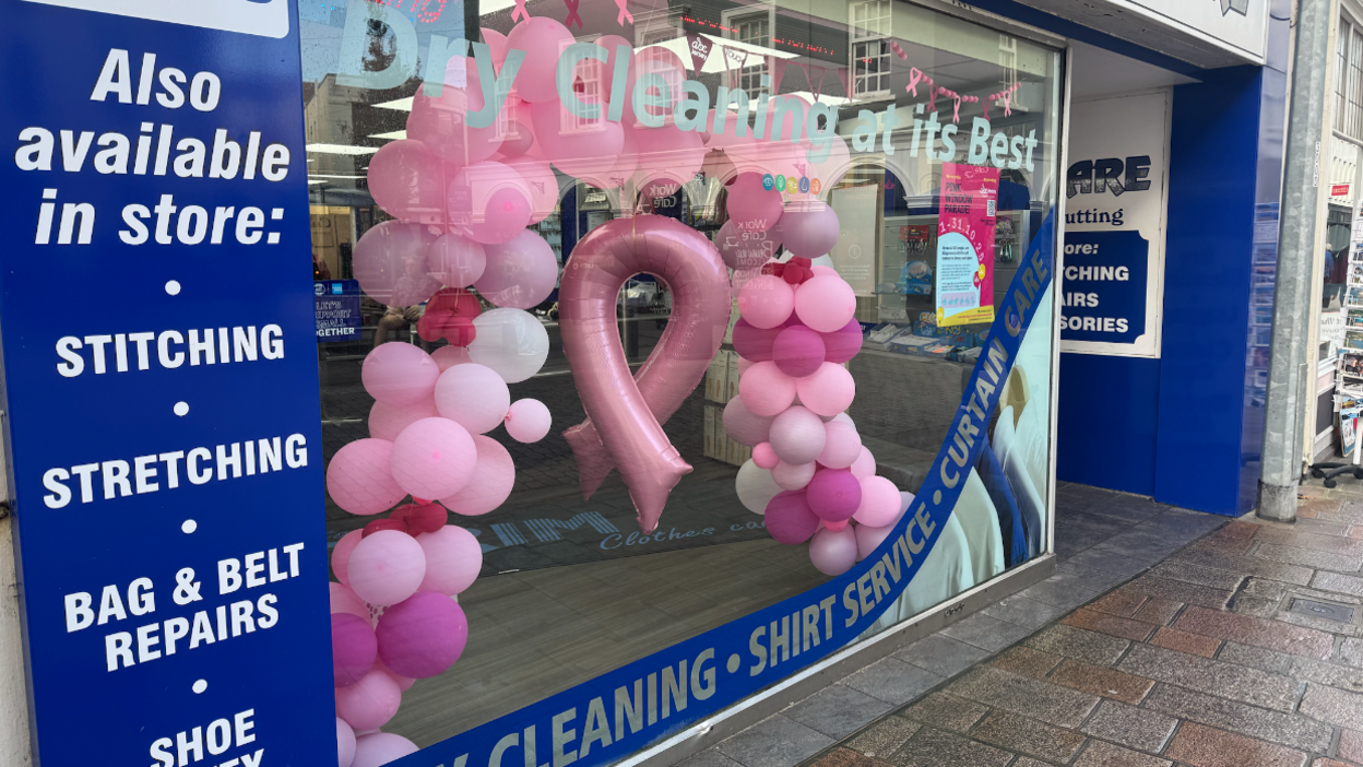 A pink balloon arch with the pink breast cancer emblem balloon in the middle in a dry cleaning company's window display.