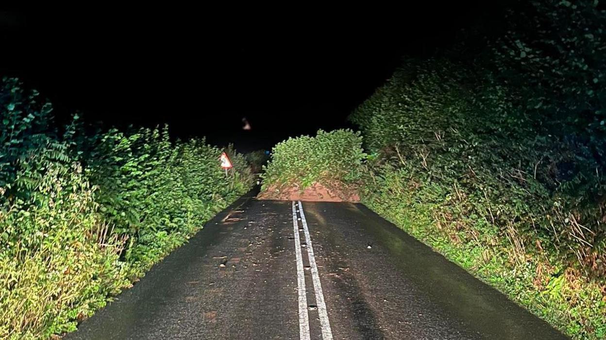 A mound of earth blocking the A379 in Modbury. It is night time and the mound also has shrubbery on it. The road looks wet following rainfall. The road and hedges either side of it have been lit up by the lights of a vehicle.