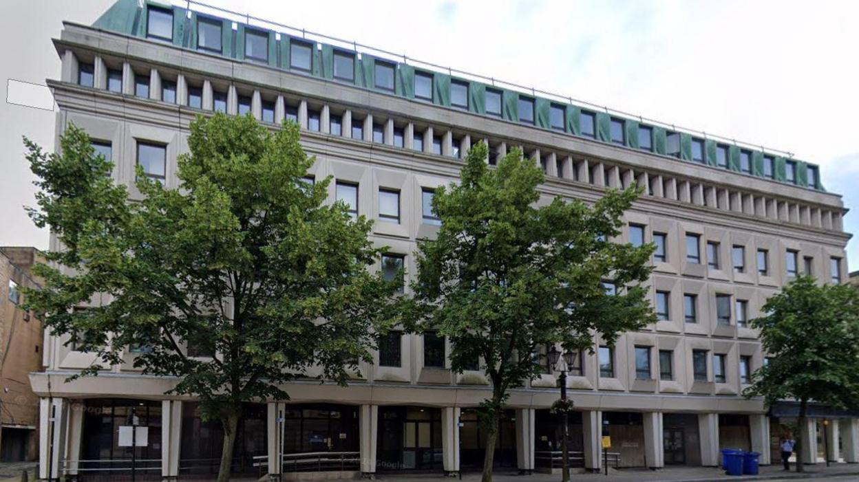 A large grey stone five storey building with rectangular windows. There are three trees lining the pavement in front.