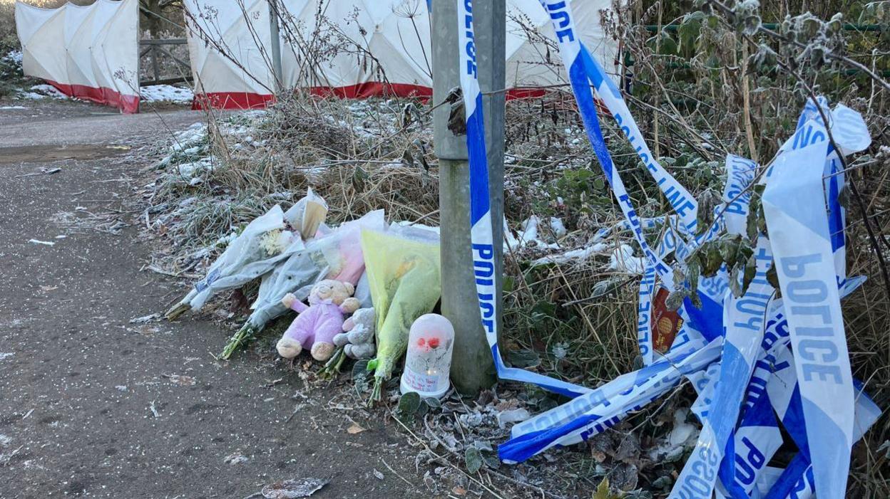 Flowers and a teddy bear rest near a lamppost, with bunched-up blue-and-white police tape lying to the side