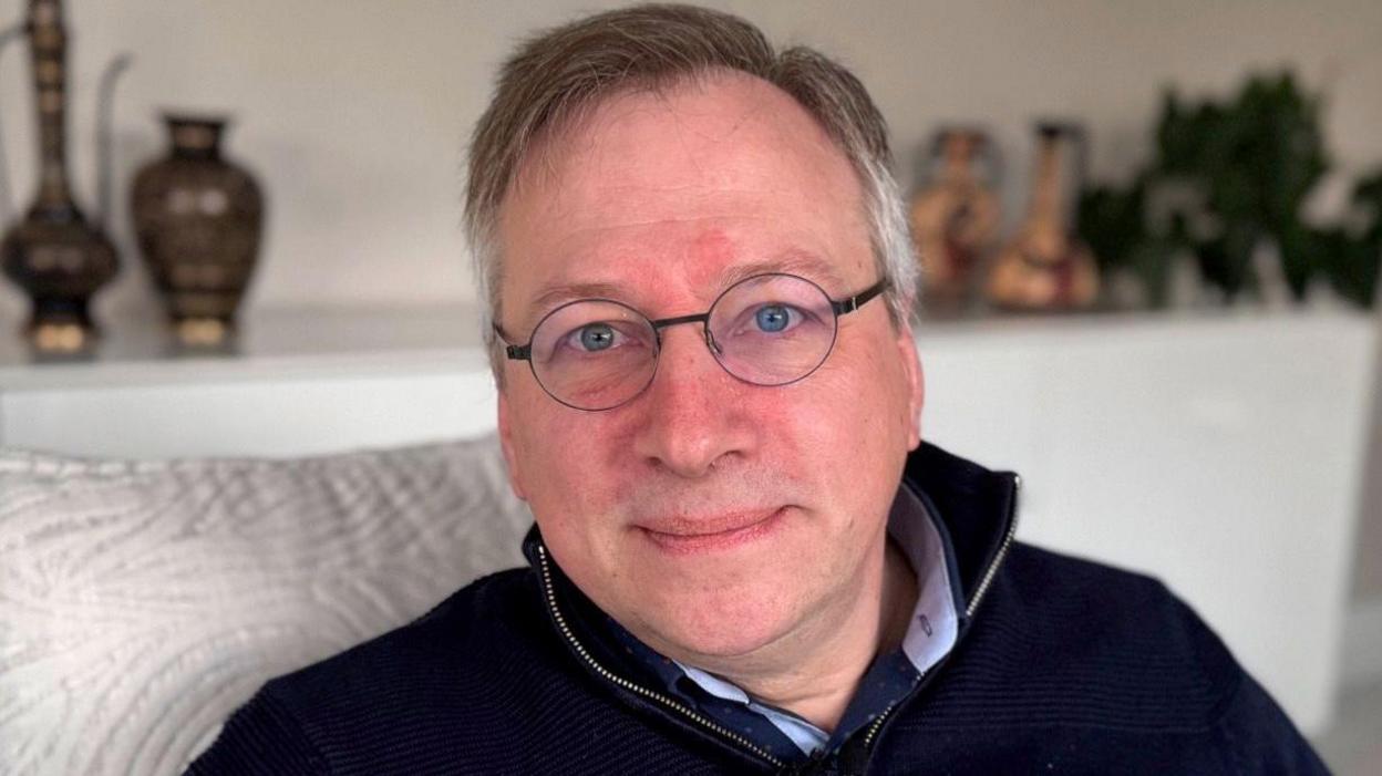 A man with round glasses and short grey hair sits on a silver sofa, looking down the lens of the camera. He has his lips pursed and his eyes are a piercing blue colour. He is wearing a navy quarter zip jumper with a silver zip.