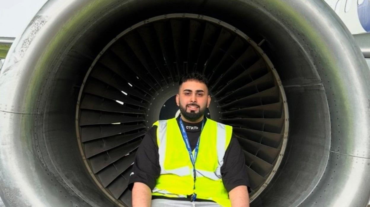 Faizan Mujtaba, with a beard, is smiling. He is sitting in the centre of the photo wearing a high-vis jacket and a lanyard.