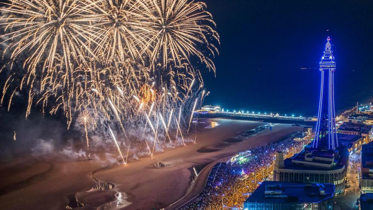 Aerial view of the fireworks display on the beach beside Blackpool's North pier with crowds of people watching on the promenade in the evening with the Blackpool Illuminations on and Blackpool Tower lit up purple in the background.