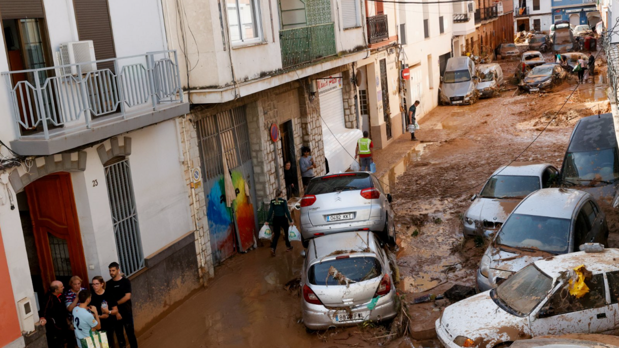 People gather in a mud-covered street with damaged cars piled on top of each other in Paiporta on 31 October 