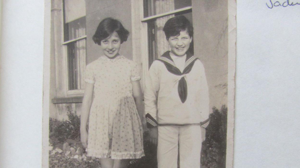 Black and white photo of a young girl in a dress and a young boy standing side by side smiling at the camera.