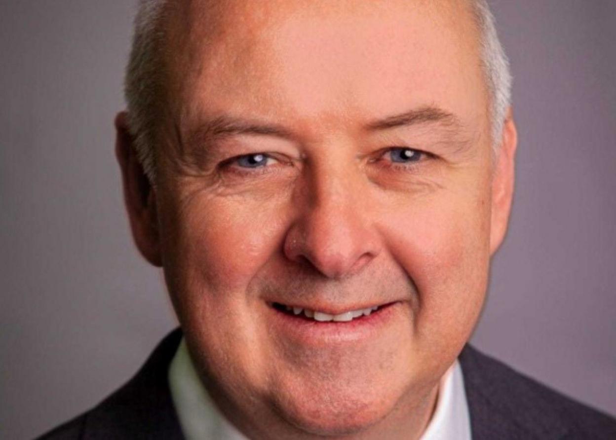 Commissioner Ben Adams is looking at the camera, smiling. He is wearing a dark suit and is in front of a purple background.