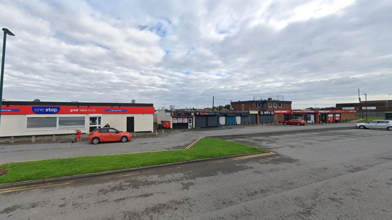 The shops off Allendale Road. An orange car is parked outside a One Stop convenience store to the left. The row of shops continue to the right and include a pizza shop, a kebab shop and a Family Shopper store.