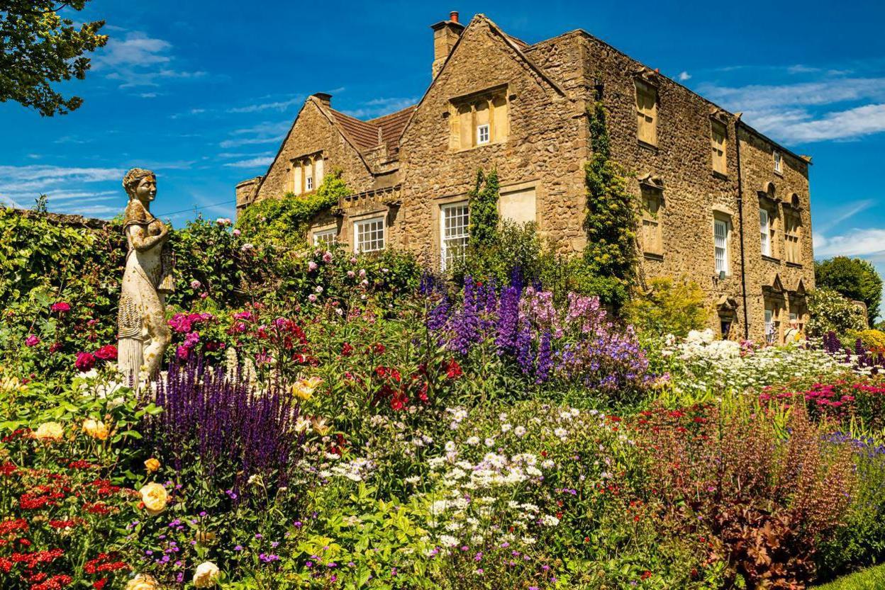 The gardens at Thornton Hall are in full bloom with roses and other pink, purple and yellow flowers. There is a statue of a woman in the midst of the flowers.