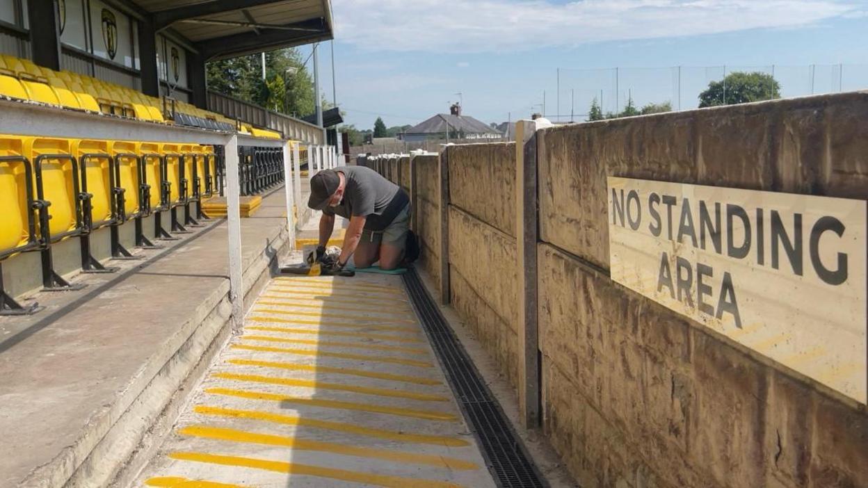 A man is on his hands and knees painting yellow lines on the concourse of a non-league stadium.