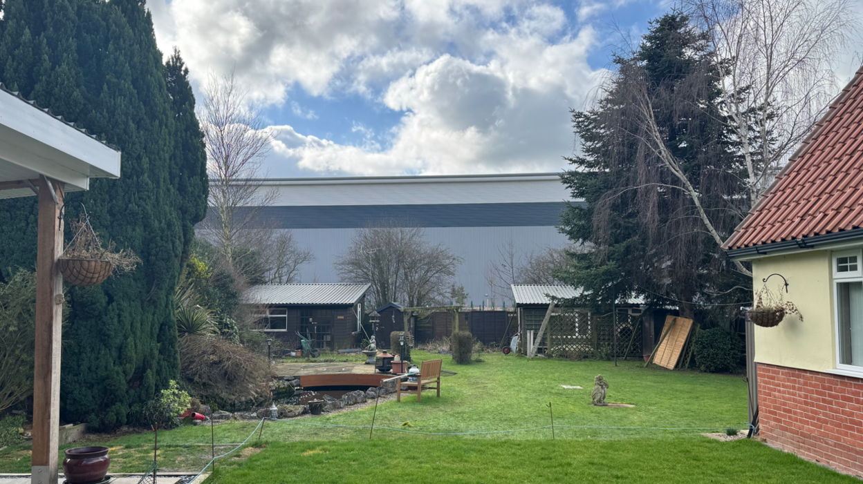 A large warehouse can be seen at the end of a big back garden, which has a small pond and multiple sheds. 