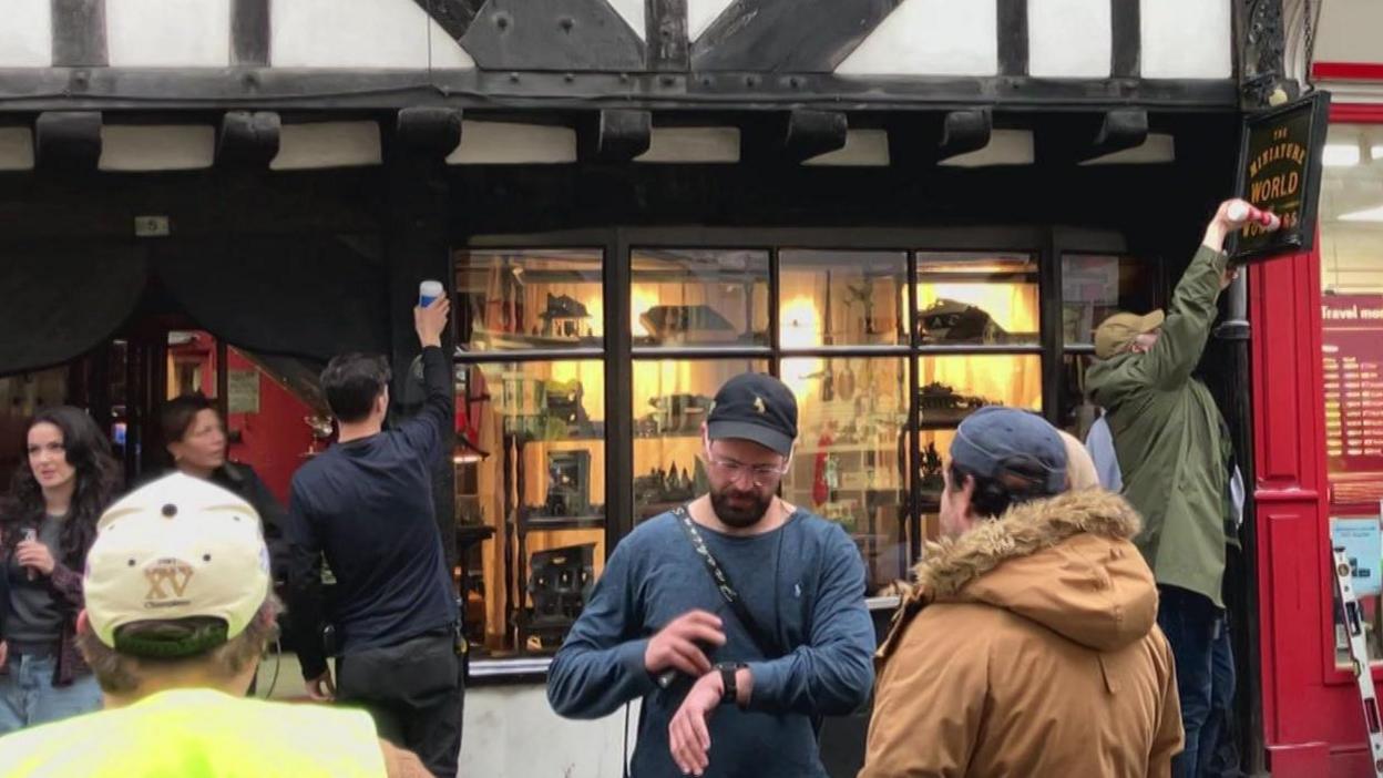 A group of people, many wearing caps, some wearing hi-vis jackets. Two people are pouring table salt from bottles onto window ledges and the sign outside a shop.