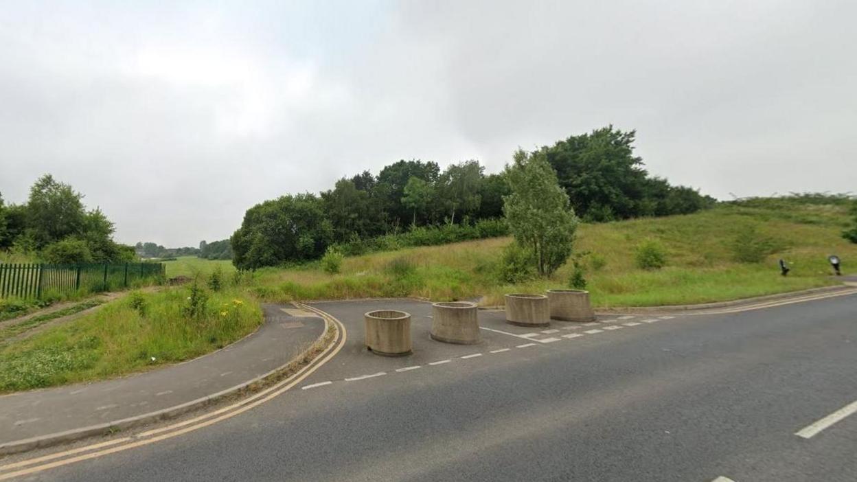 Land earmarked for development, with grass, trees and four bollards blocking a road 