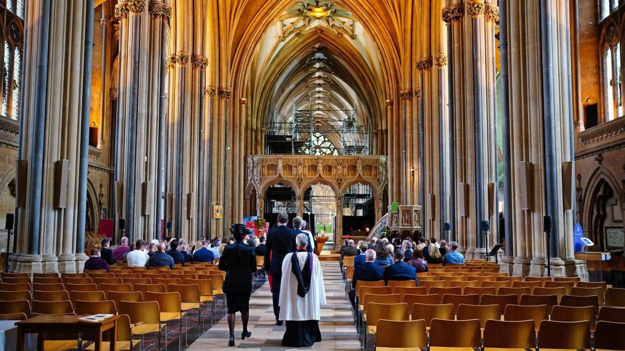 A view looking down the main central aisle of Bristol Cathedral with people sitting either side and a member of the clergy walking in robes walking away from the camera. The picture is taken during a memorial service for  British aid worker James Kirby.