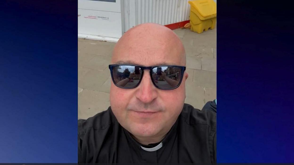 Father Christopher Woods filming his video near the Botley Road development. He is wearing sunglasses.