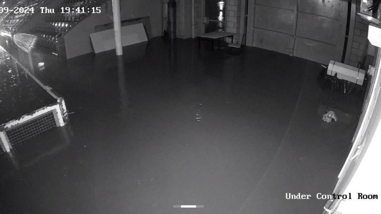 A black and white security footage screengrab. There is floodwater next to football stands