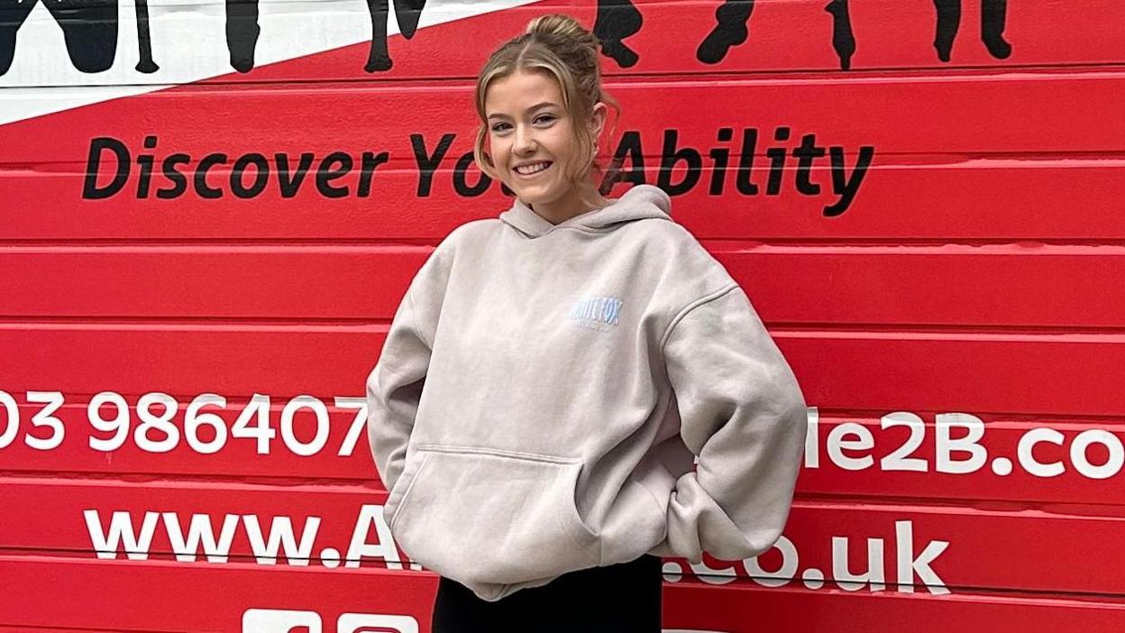 Emma Daniels, who has blonde hair, is looking at the camera and smiling. She is wearing an off-white coloured hoodie and black trousers. She is stood in front of a red door at Able 