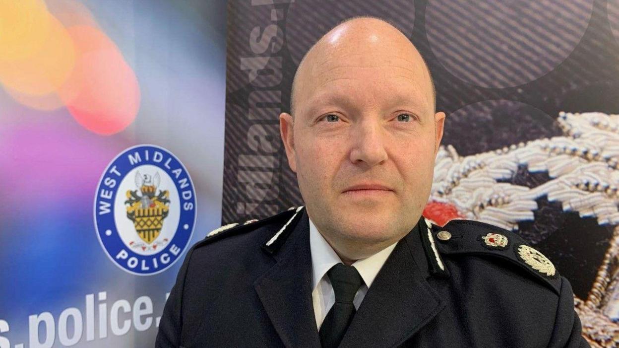 A man in a police uniform, who is bald, stands in front of a poster. The uniform consists of a black jacket, a white shirt and a black tie. On the poster behind him, a blue and white logo with the words "West Midlands Police" can be seen.
