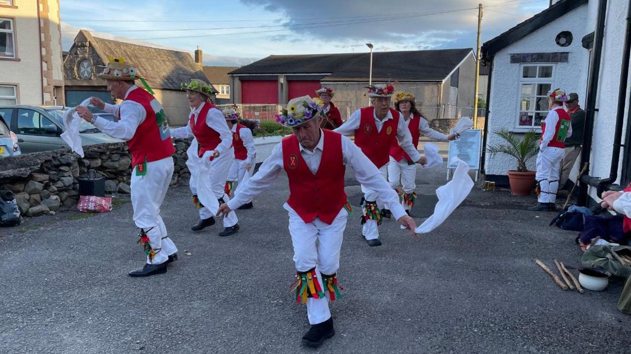 Members of Furness Morris Dance dancing in red and white clothing in Ulverston in May 2022. there are about eight of them wearing hats with colourful artificial flowers at the brim.
