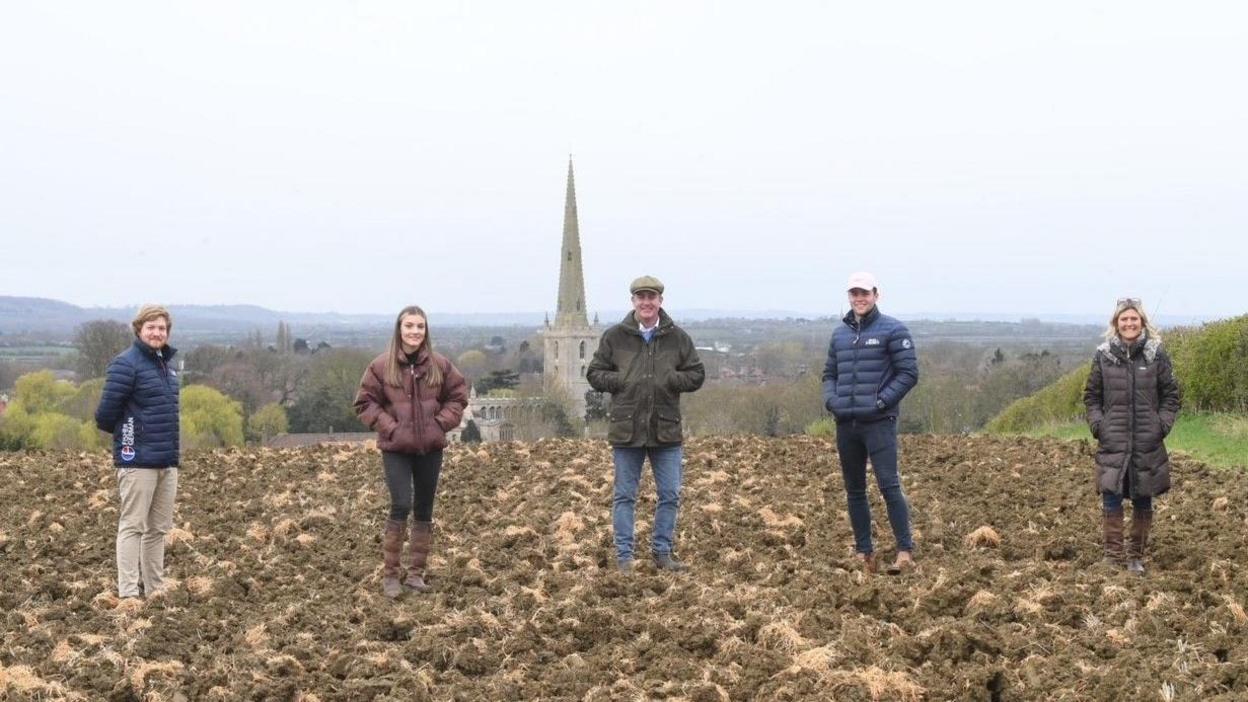 An image of five people stood in a line on a farm with a church steeple in the background
