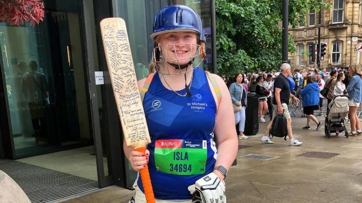 Isla Bayliss is wearing cricket gear, including a blue helmet and white pads, and holding a cricket bat with extensive writing on it in her right hand.