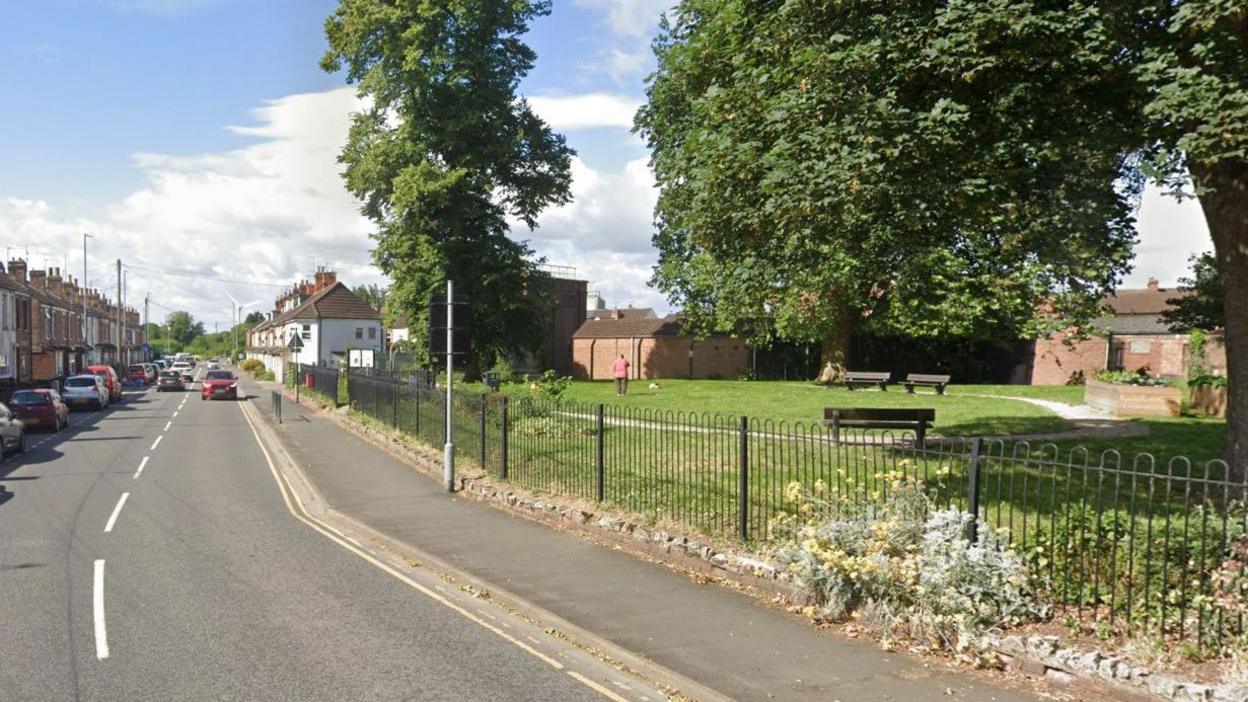 Terraced houses line a road beside a small park, with green grass, benches and trees, in Ashcroft Road, Gainsborough