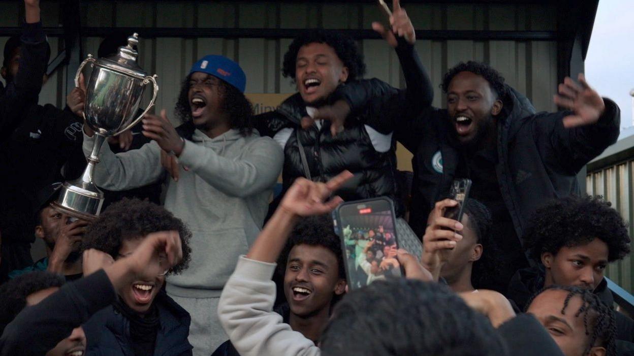 A silver metal cup is waved in the air by a fan surrounded by other fans on what appears to be part of a football terrace, with metal sheets at the back behind them. The fan with the cup has a blue cap on backwards, a grey top and grey trousers while the other fans have puffy black jackets, some hold phones aloft as if recording the scene.