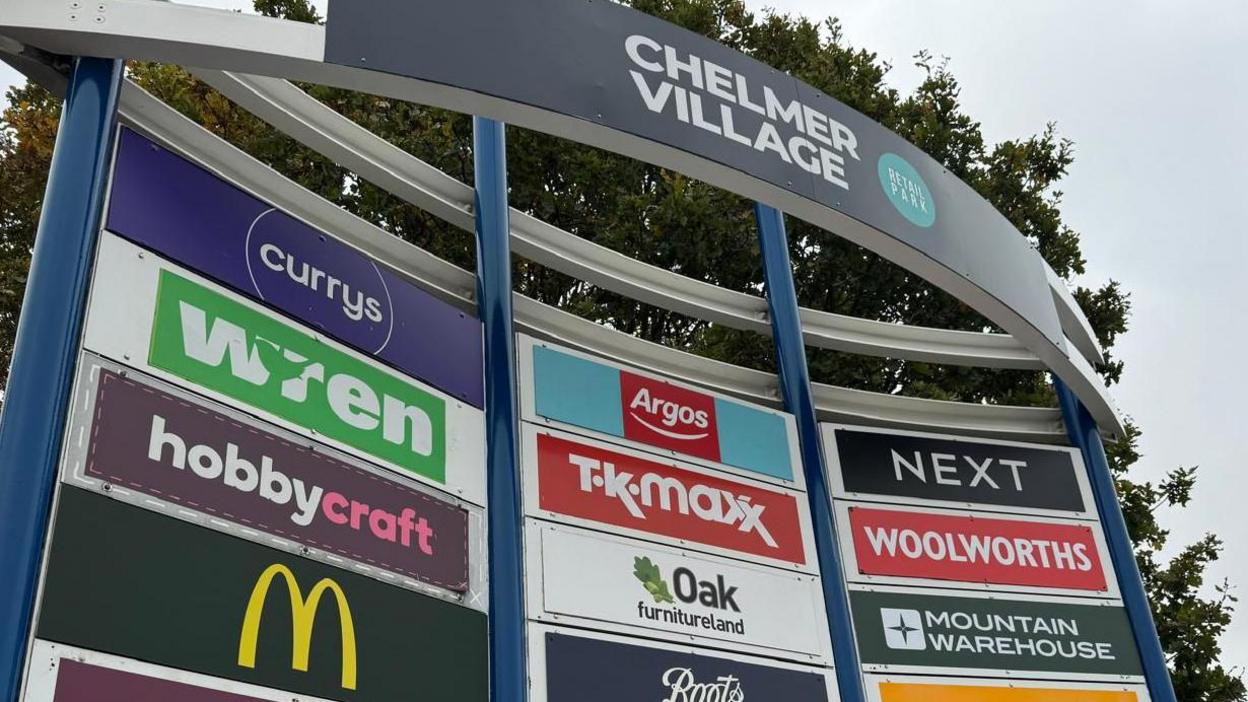 A sign for Chelmer Village that lists all the brands within the shopping park, including McDonald's, Currys and Argos. A red Woolworths sign has also been placed on the board on the right hand side. It is underneath a sign for Next and above one for Mountain Warehouse. 