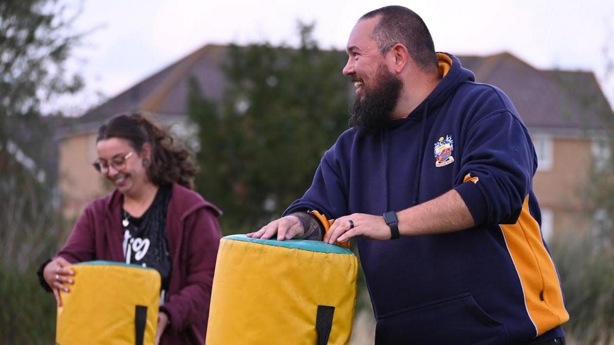 Mr Goacher pictured during a practise with the rugby bags, smiling off to the left of the image. He is wearing a navy and yellow hoodie with a logo on its lapel. On the left side of him, a woman in a red hoodie is holding another rugby bag and smiling.