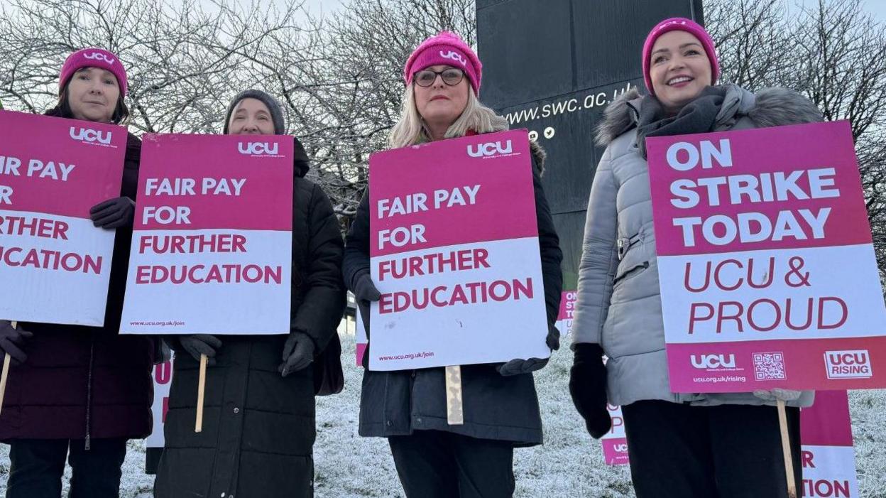Four people are standing in front of a sign for South West College. There is snow on the ground behind them and they are all wearing hats and carrying placards from the UCU calling for Fair Pay for Further Education